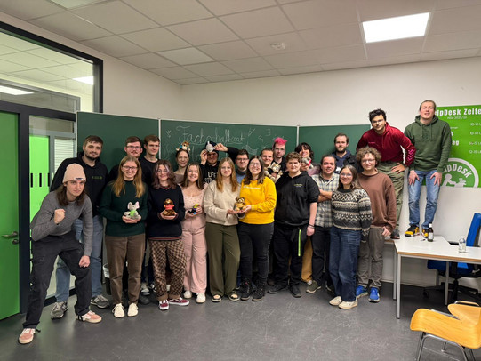 Ein Gruppenfoto des Fachschaftsrats in einem Seminarraum. Die Mitglieder posieren gut gelaunt vor einer grünen Tafel, auf der "Fachschaftsrat" und mathematische Formeln stehen. Einige halten kleine Plüschtiere in den Händen.